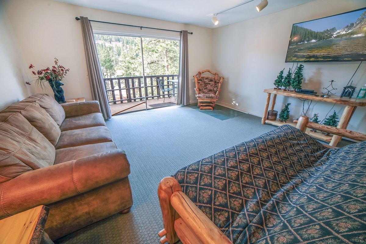 A cozy living space features a spacious sofa and a wooden rocking chair, positioned for optimal viewing of the large wall-mounted television. A sliding balcony door opens to a scenic view of the surrounding natural landscape, allowing ample light into the room.