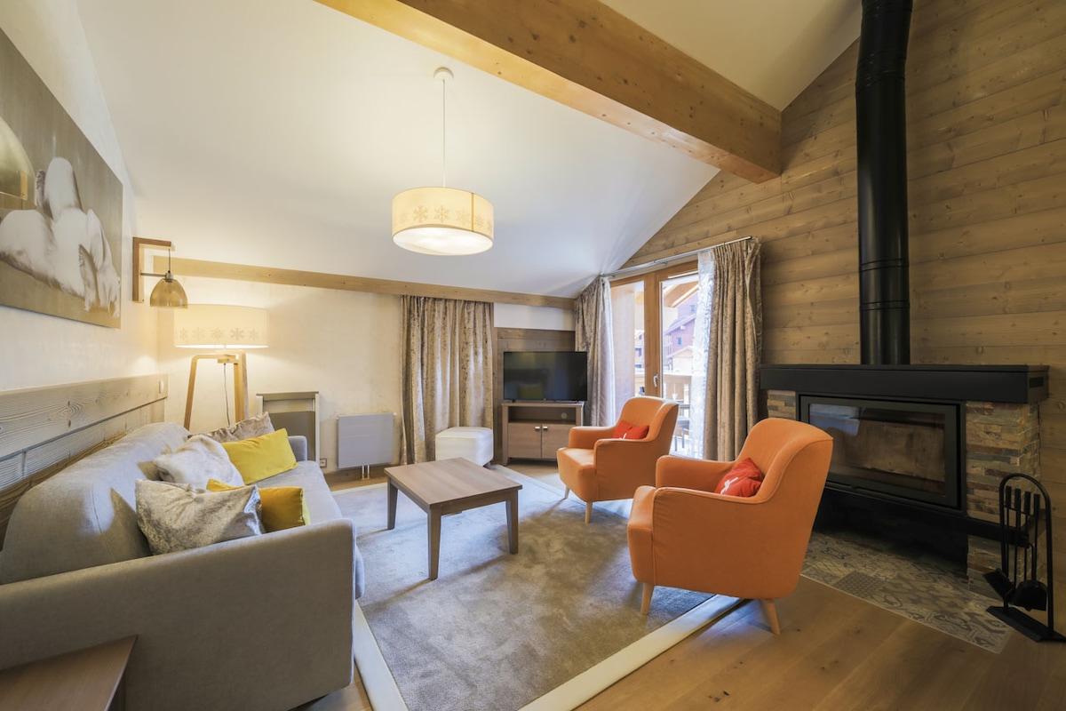 The living room showcases a spacious sofa accompanied by two armchairs, all positioned around a wooden coffee table. Warm wood paneling adds texture, while light flows in through large windows. A modern light fixture hangs overhead, and a fireplace is visible in the corner.