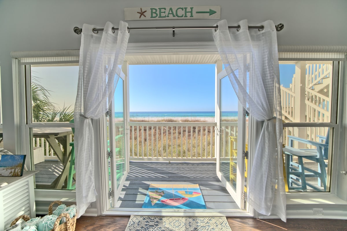 Large glass doors open to reveal expansive ocean views, allowing natural light to fill the space. A cozy area rug adds texture to the wooden floor, while sheer white curtains gently frame the scene. The balcony outside is accessible for enjoying the beachfront scenery.