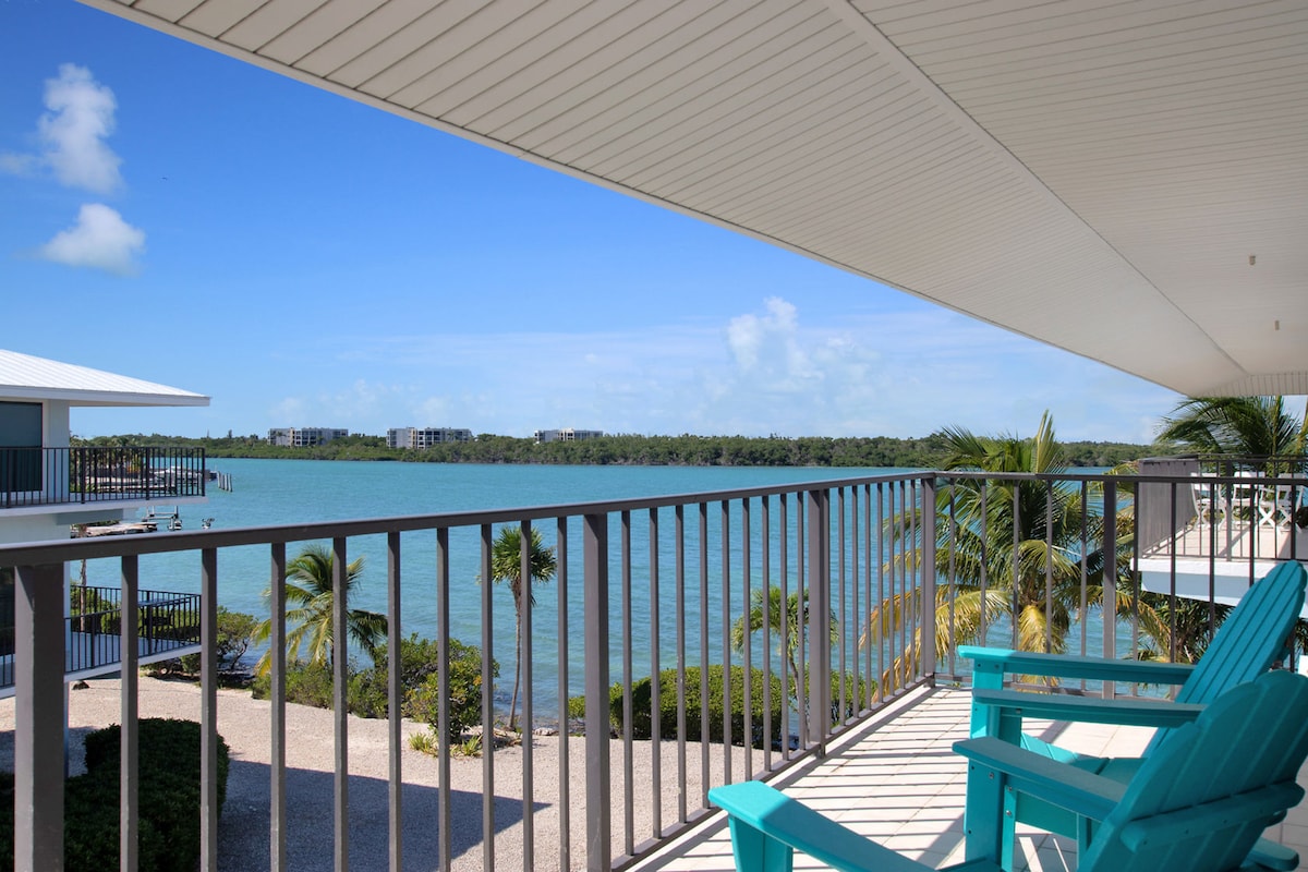 A balcony view overlooks calm waters with lush greenery lining the shore. Two bright turquoise chairs invite relaxation, while a gentle breeze can be sensed through the open space. The sky is clear, providing a vivid contrast to the serene surroundings.