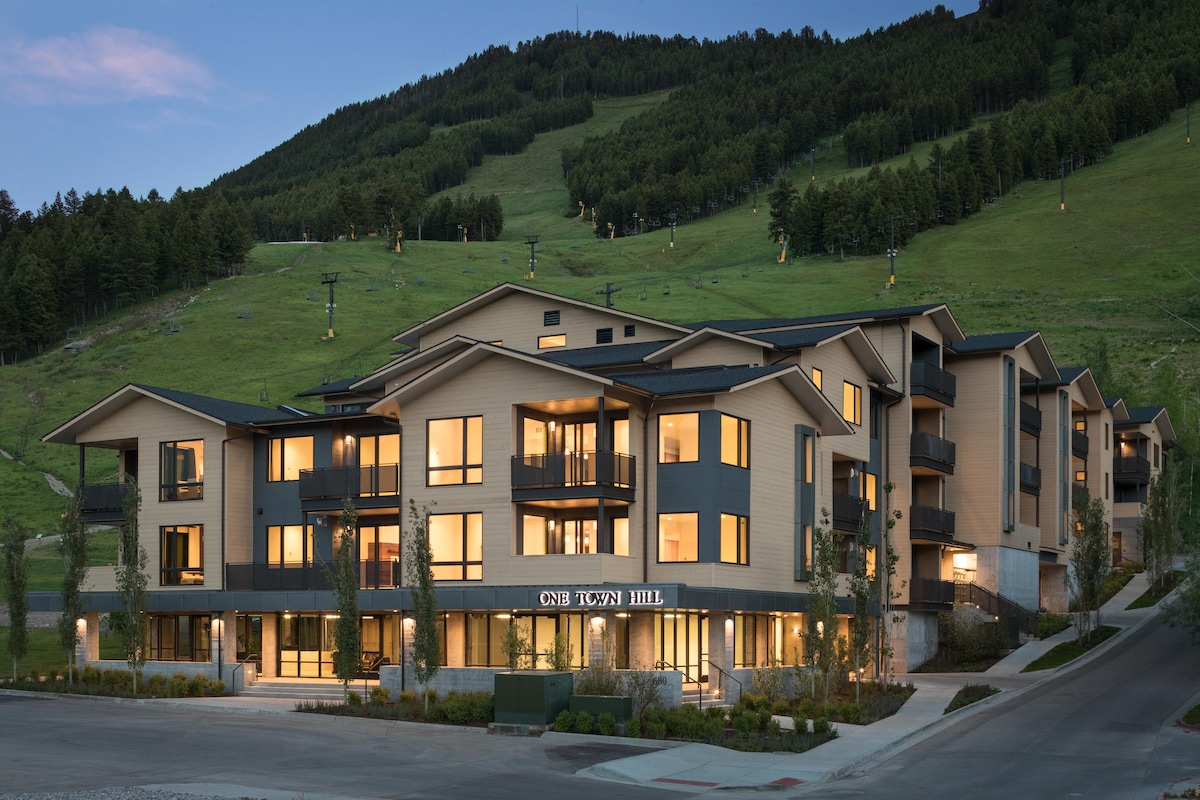 The exterior of One Town Hill showcases a modern architectural design, complemented by surrounding green hills. The building features multiple levels with balconies and large windows, allowing natural light to fill the interior spaces. Pathways and landscaped areas enhance the welcoming appearance.