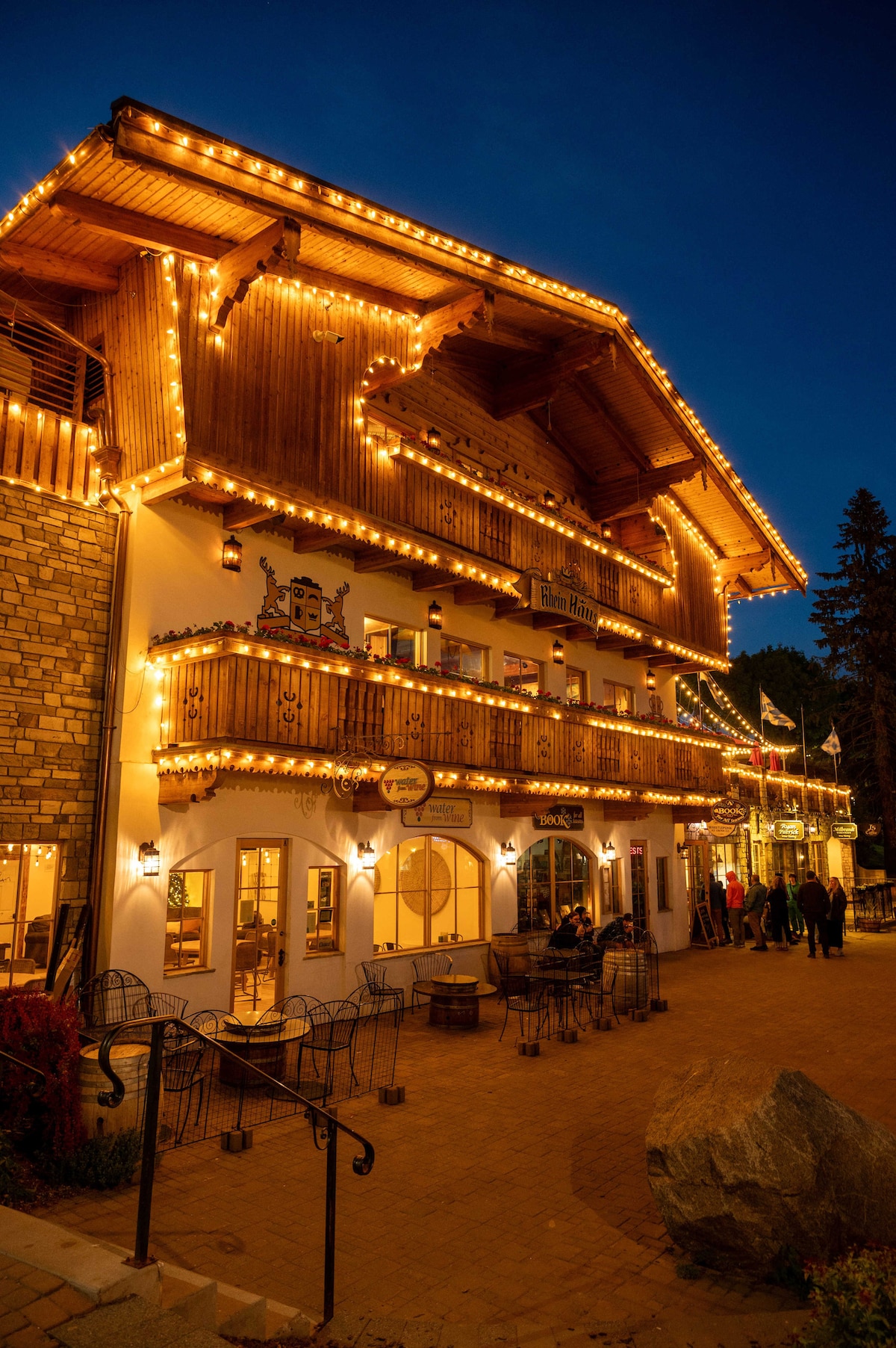 The exterior of a charming building is adorned with warm, glowing lights, creating a festive ambiance. A spacious balcony showcases decorative elements, while seating areas are arranged along the walkway. The scene is set at dusk, with a clear view of the inviting structure and nearby visitors.