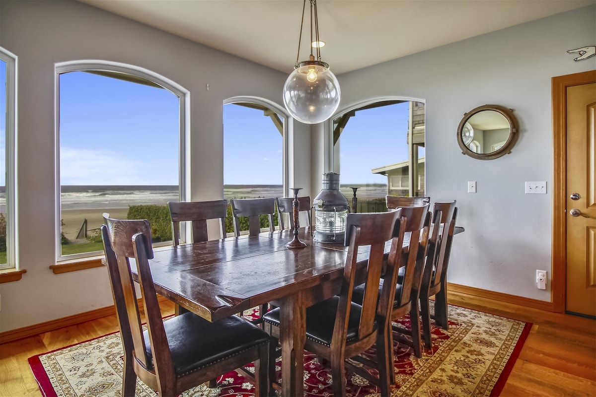 A spacious dining area features a large wooden table set for eight, surrounded by high-backed chairs. Large windows provide expansive views of the beach, allowing natural light to fill the room. A pendant light hangs above the table, complemented by a decorative mirror on the wall.