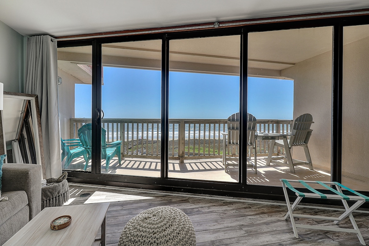 Large glass sliding doors offer a panoramic view of the ocean from the living space. A spacious balcony is visible, furnished with two chairs and a chaise lounge. Natural light fills the room, while comfortable seating is arranged along a neutral-colored wall.