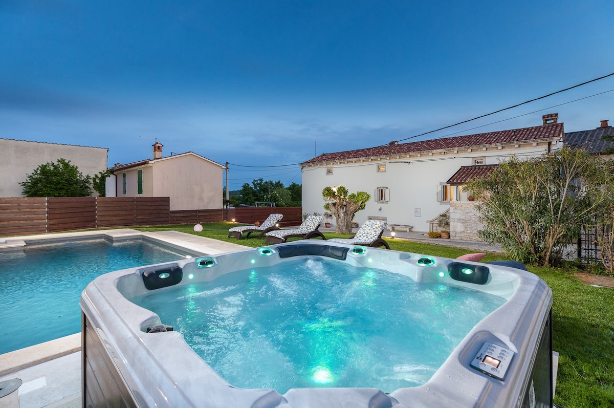 A private spa hot tub is positioned near a swimming pool within a spacious garden, offering a tranquil retreat. The modern hot tub features LED lighting, and lounge chairs are arranged nearby, surrounded by lush greenery and the charming architecture of the villa in the background.