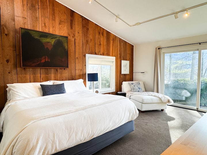 Sunlit bedroom with king bed, wood accent wall, chaise lounge, and patio views