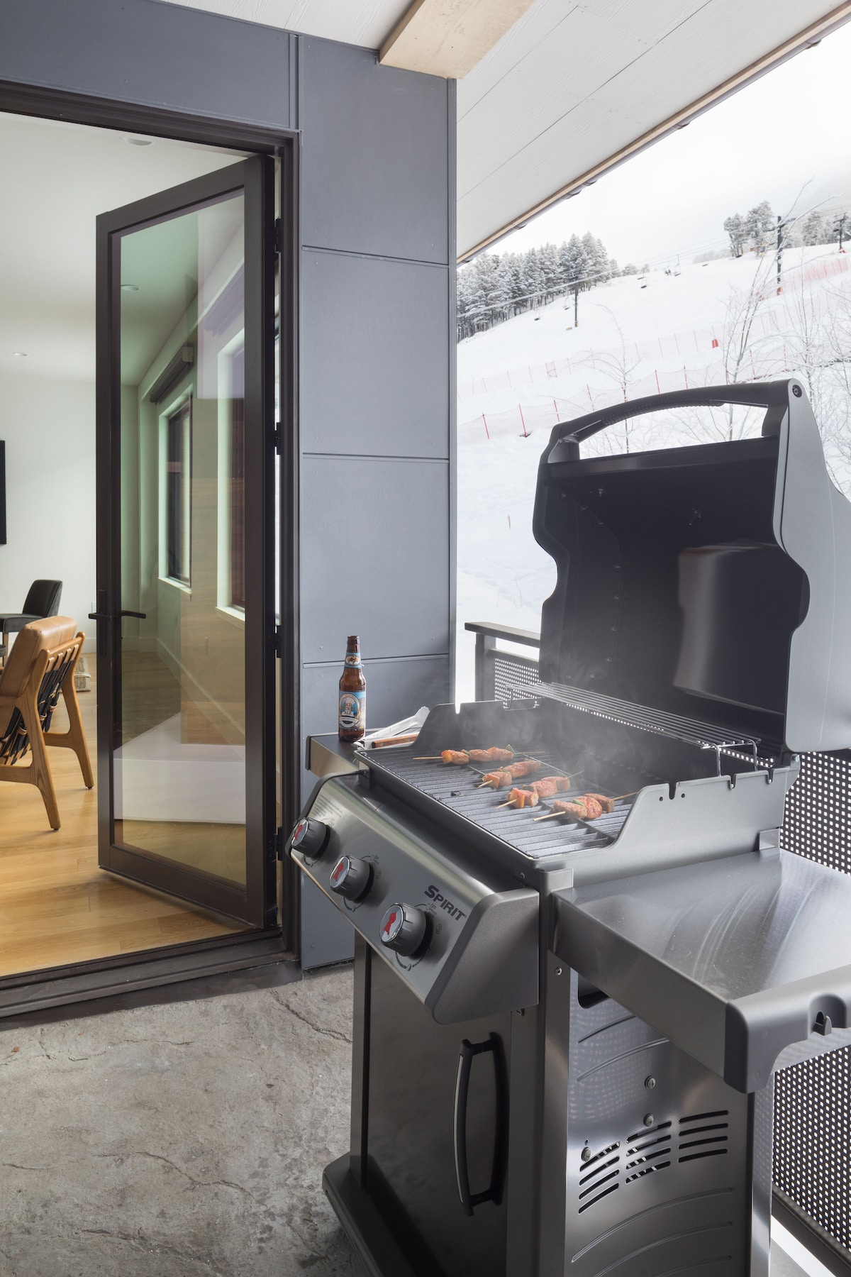 A modern grill is positioned on a balcony, with smoke rising from the cooking surface. An open door reveals the interior living space, while snow-covered slopes are visible in the background, highlighting the mountainous surroundings.