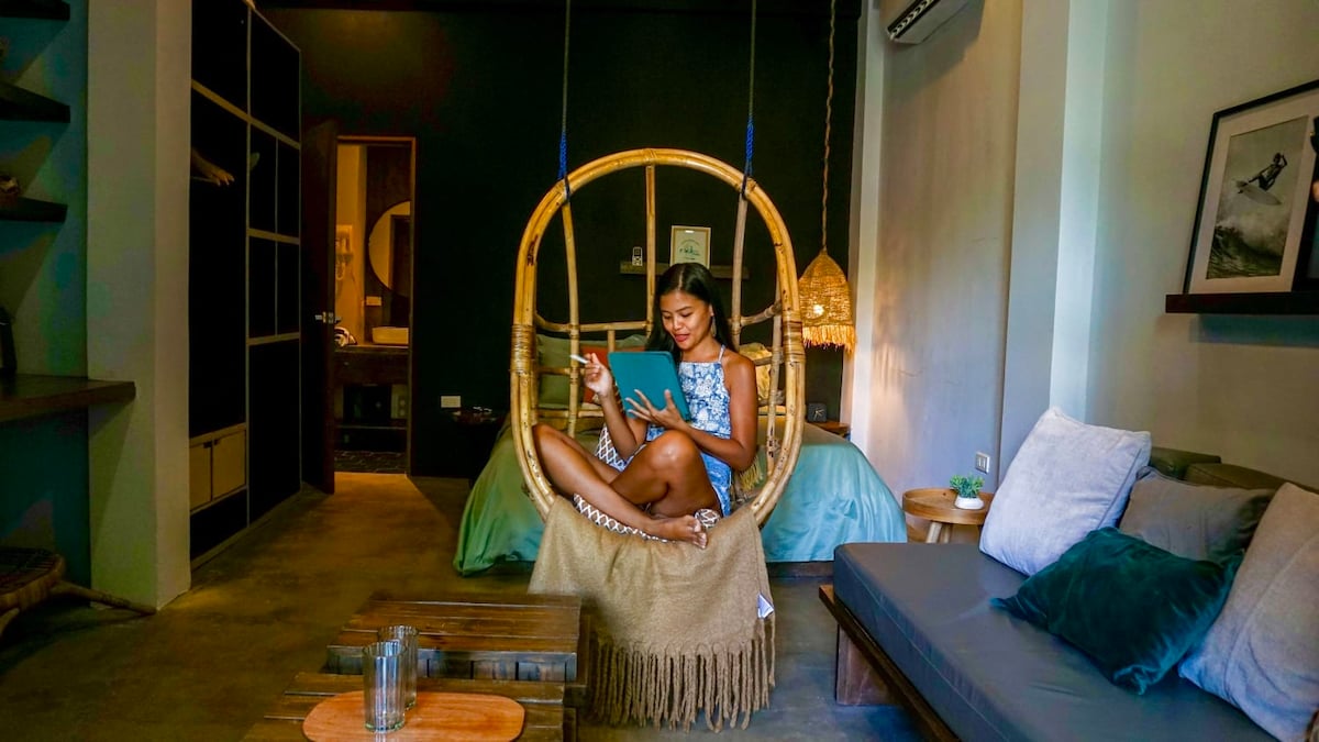 A cozy interior features a unique hanging chair with a textured beige cushion, providing a relaxed space for reading. A spacious bed with light bedding is set against a dark accent wall, while a neutral-toned sofa adds to the inviting ambiance.