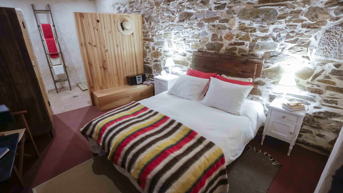 The room features a spacious bed with white linens and striped blankets. Exposed stone walls add character, complemented by wooden elements. Two bedside tables with lamps provide soft lighting. A wooden wardrobe and a rustic ladder can also be seen, enhancing the room's inviting atmosphere.