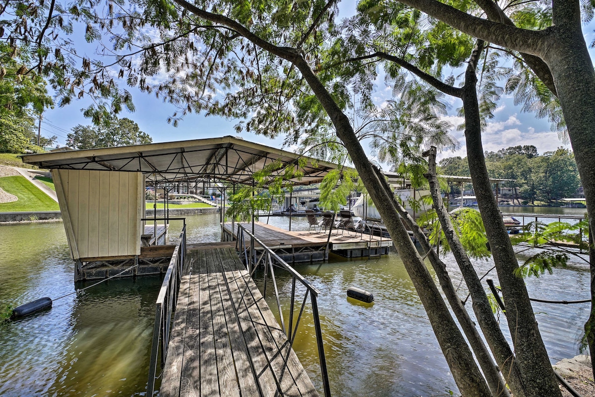 A private dock is shown extending over the water, featuring a covered area for shade. Surrounding trees provide natural framing, and dock space is visible for multiple boats, reflecting the waterfront setting and serene atmosphere of the location.