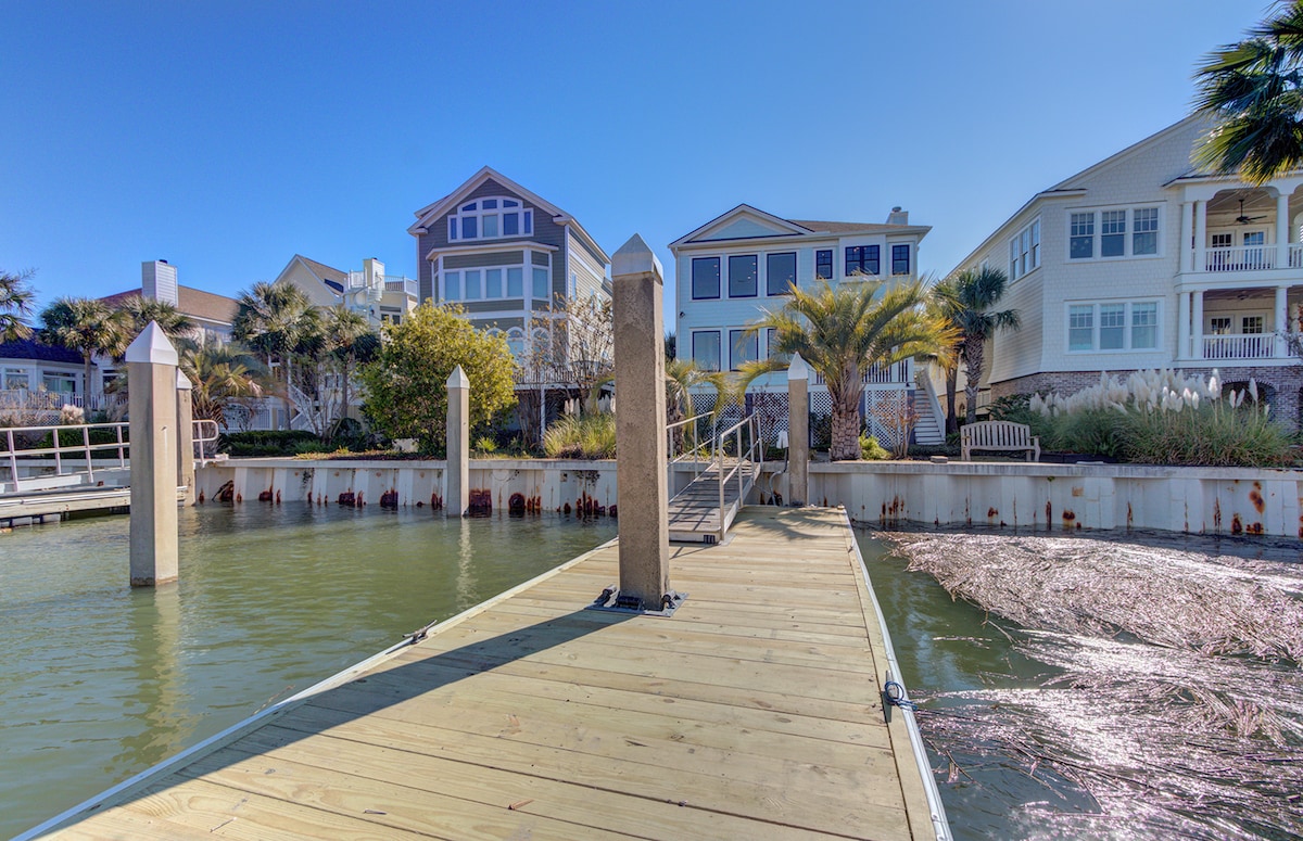 A wooden pier extends from the water towards several waterfront homes. Surrounding vegetation includes palm trees and grasses, creating a natural backdrop. The clear blue sky reflects off the calm water, highlighting the serene setting of this coastal community.