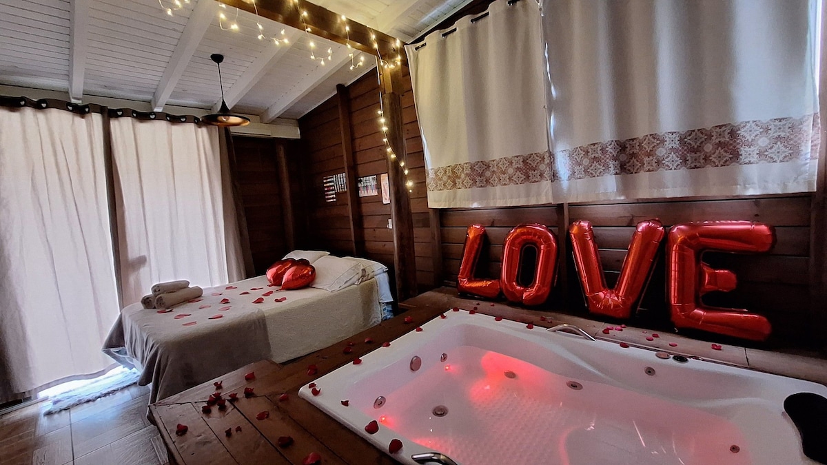 An intimate room featuring a spacious bed adorned with rose petals. A bubble-filled Jacuzzi is lit with soft red light, while large red letters form the word 'LOVE' against the wooden wall. Natural light filters through sheer curtains, creating a warm ambiance.