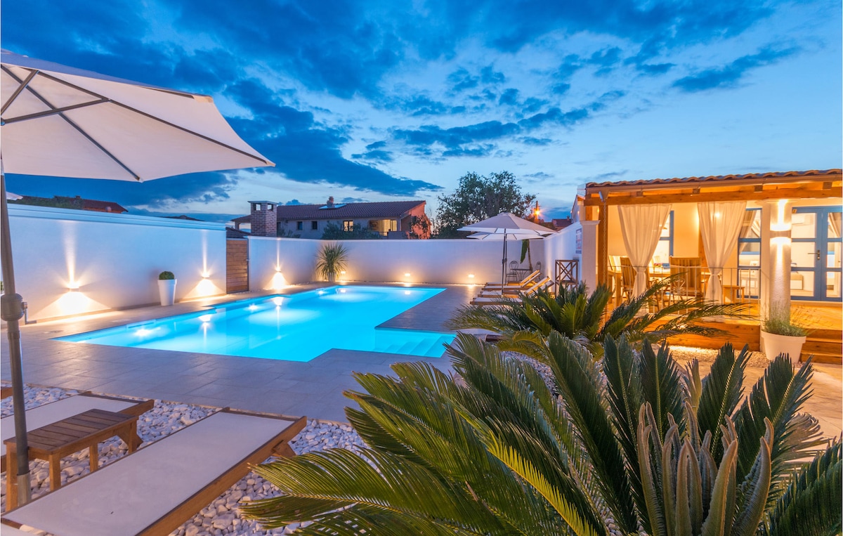 A tranquil outdoor area features a swimming pool illuminated by soft lighting, surrounded by palm plants and sun loungers. An umbrella provides shade, while a cozy seating area with draped curtains offers space for relaxation. The sky is painted in shades of blue as evening sets in.