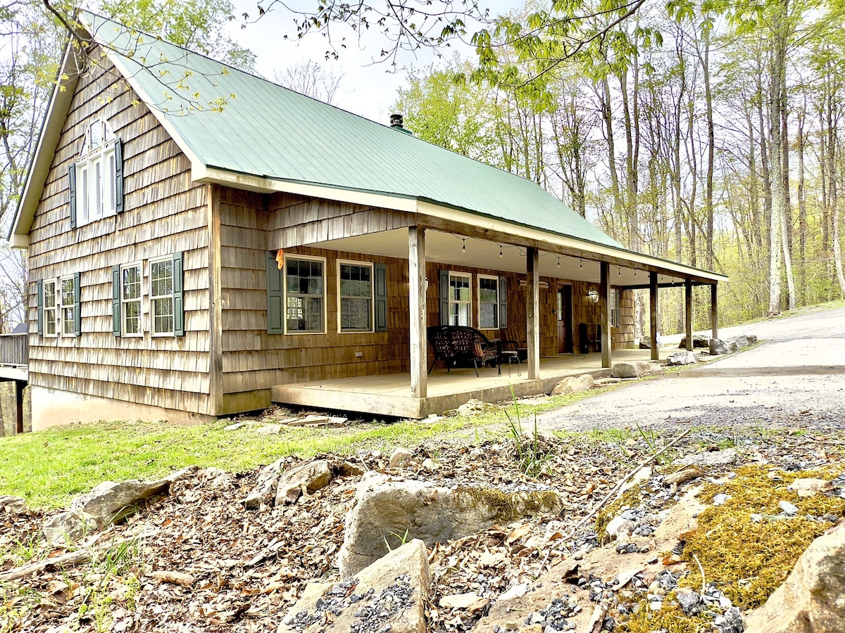 A charming mountain retreat is featured, showcasing a spacious front porch with a swing. The exterior is adorned with wooden shingles and a green metal roof. Surrounding nature provides a serene backdrop, while a well-maintained driveway leads to the entrance.