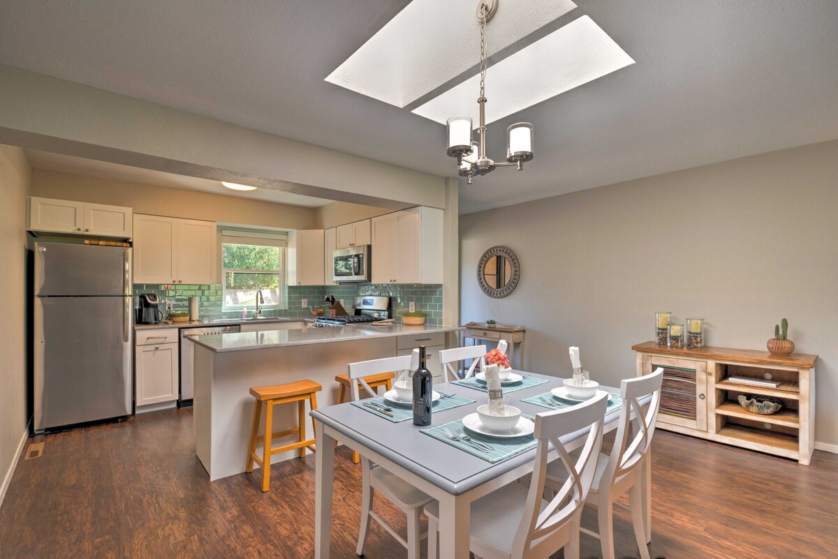 The kitchen area features modern stainless steel appliances, including a refrigerator and oven. A white dining table is set for four, complemented by light blue table settings. Natural light enters through a skylight, enhancing the spacious open layout that connects to the living area.
