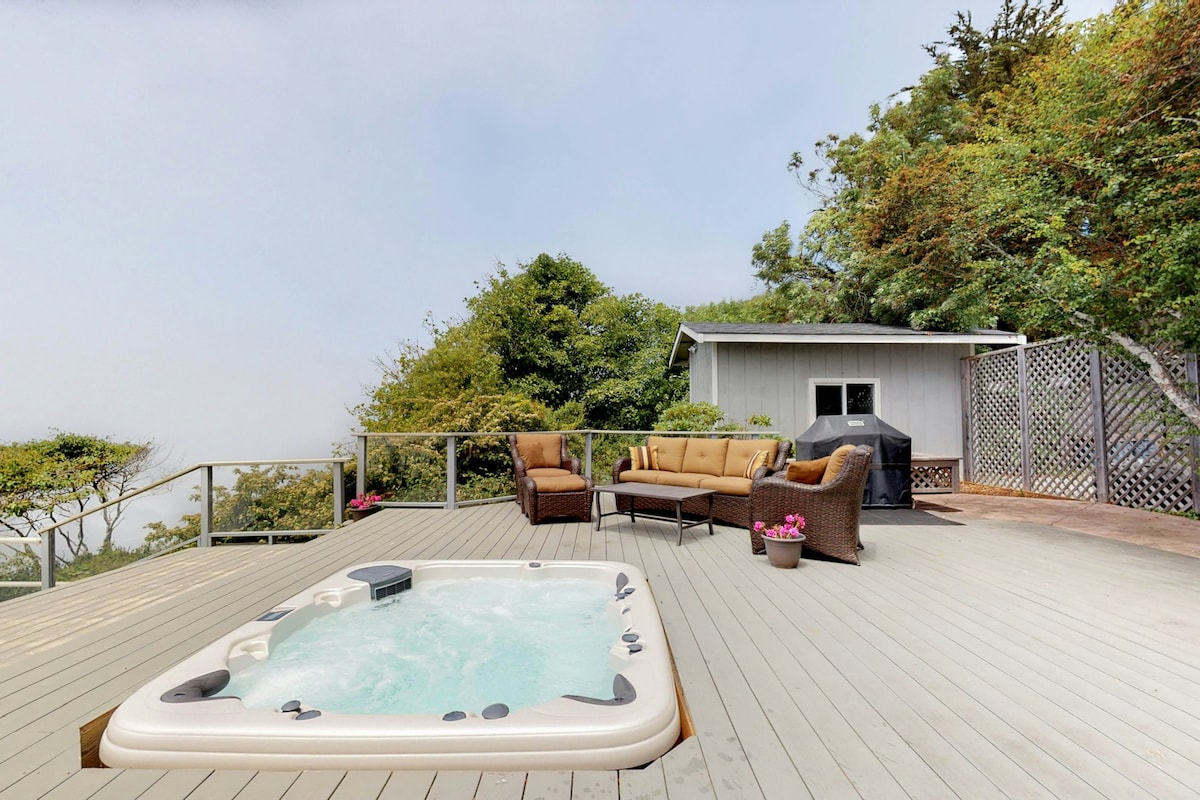 A spacious outdoor deck features a sunken hot tub filled with water. Nearby, a seating area includes a sofa and chairs arranged around a coffee table. A barbecue grill is placed in the corner, surrounded by greenery and potted flowers.