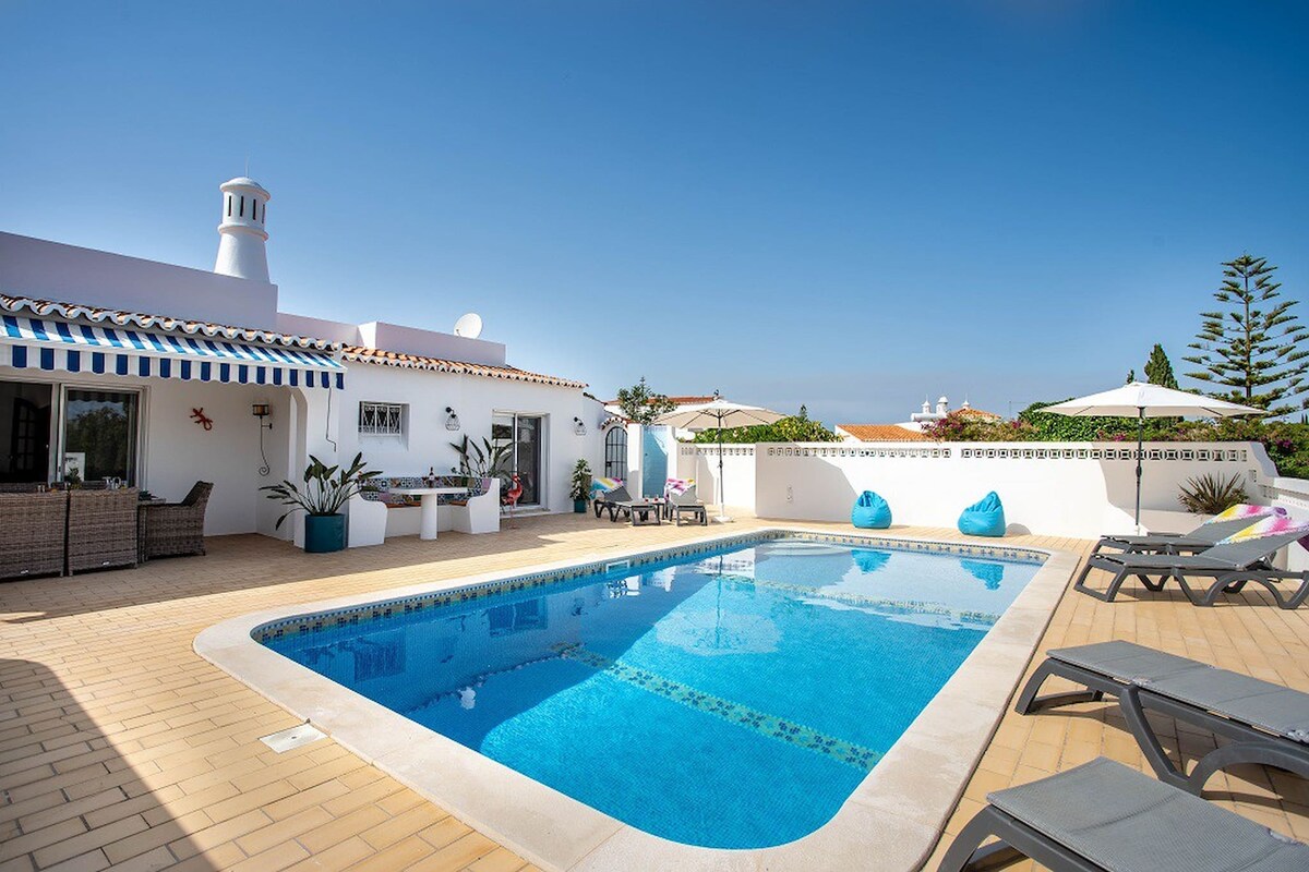 A spacious outdoor area features a sparkling swimming pool surrounded by sun loungers and bean bag chairs. A patio with dining furniture is visible, and decorative plants add a touch of greenery. The clear blue sky enhances the sense of openness in the setting.