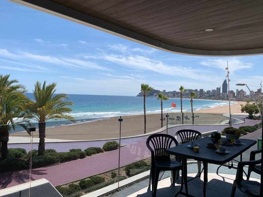 A spacious terrace is equipped with outdoor furniture, offering views of the beach and ocean. Tall palm trees line the shoreline, and the sandy beach extends along the coast. The distant skyline of Benidorm is visible under a clear blue sky.