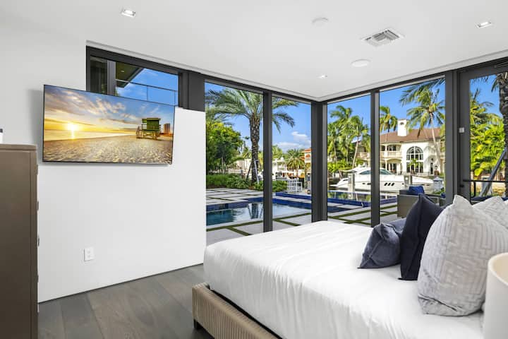 غرفة نوم مضاءة بأشعة الشمس مع زجاج ملفوف وتلفزيون ذكي وإطلالات رائعة على المياه محاطة بالنخيل