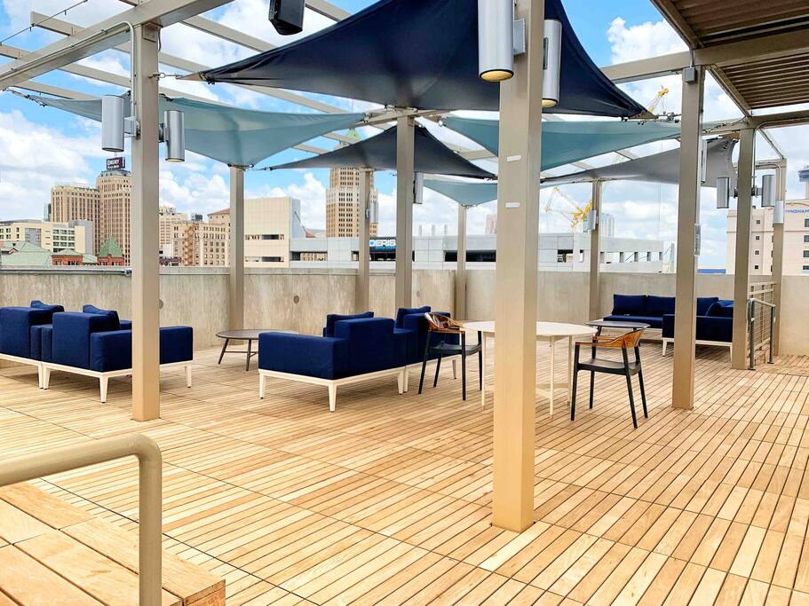 An outdoor lounge area features modern blue sofas and sleek wooden tables under triangular shades. The wooden deck is spacious, offering panoramic city views. Large windows of nearby buildings reflect a clear sky, creating a relaxed atmosphere for social gatherings.