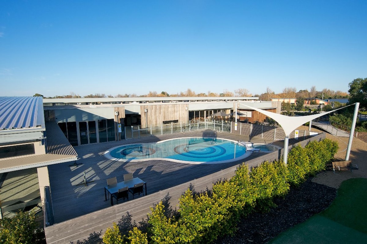 A large outdoor pool is set within a modern recreational area, surrounded by wooden decking and landscaped greenery. Sun loungers are placed nearby, and the building’s architecture features extensive glass panels, allowing natural light to fill the space.