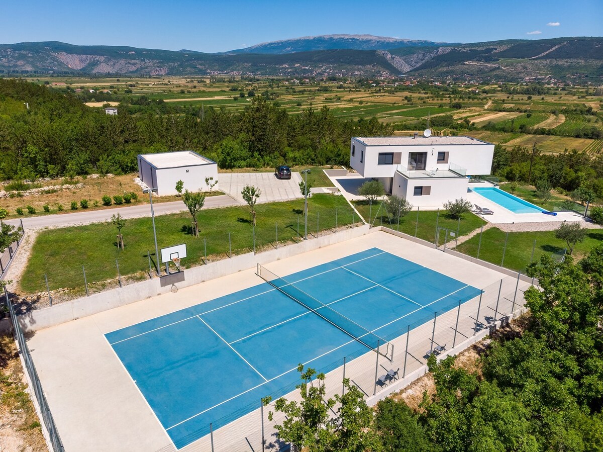 A private tennis court is visible in the foreground, bordered by a fence. A blue surface contrasts with the surrounding greenery. The villa stands in the background, spacious and modern, accompanied by a heated pool and views of the expansive valley and mountains beyond.