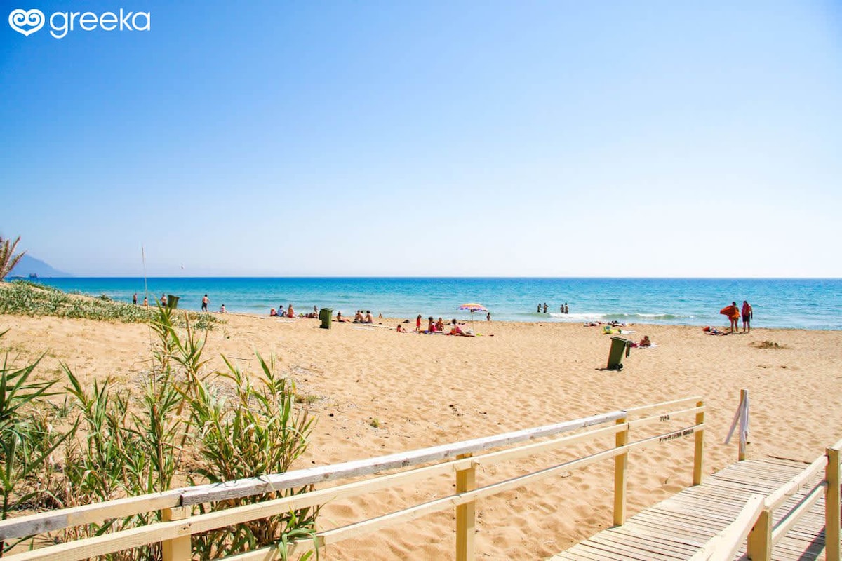 A sandy beach stretches towards a clear blue sea, populated by sunbathers and umbrellas. A wooden walkway leads from the grassy area to the shoreline, where gentle waves create a serene atmosphere. A distant horizon blends the sky and water harmoniously.