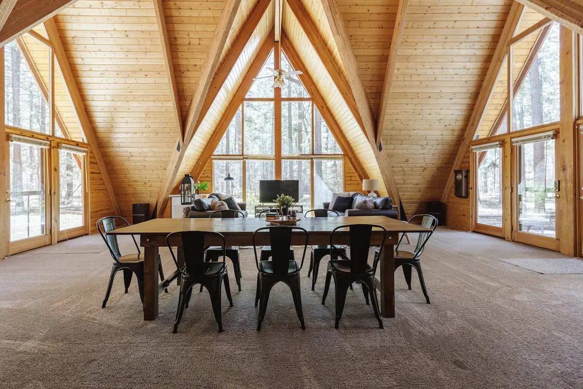 An expansive living area features a high A-frame ceiling with wooden beams, surrounded by large windows that invite natural light. A long dining table with black chairs occupies the center, while comfortable seating is arranged nearby, offering a welcoming space for gatherings.