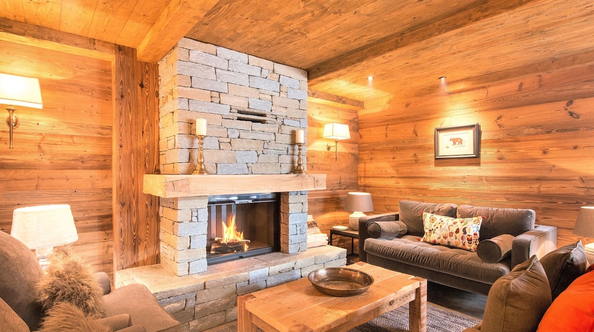 A cozy living area features a stone fireplace, providing warmth and a focal point. Soft couches are arranged around a wooden coffee table, with soft lighting from lamps illuminating the wooden walls. A decorative pillow adds a touch of color on one of the couches.