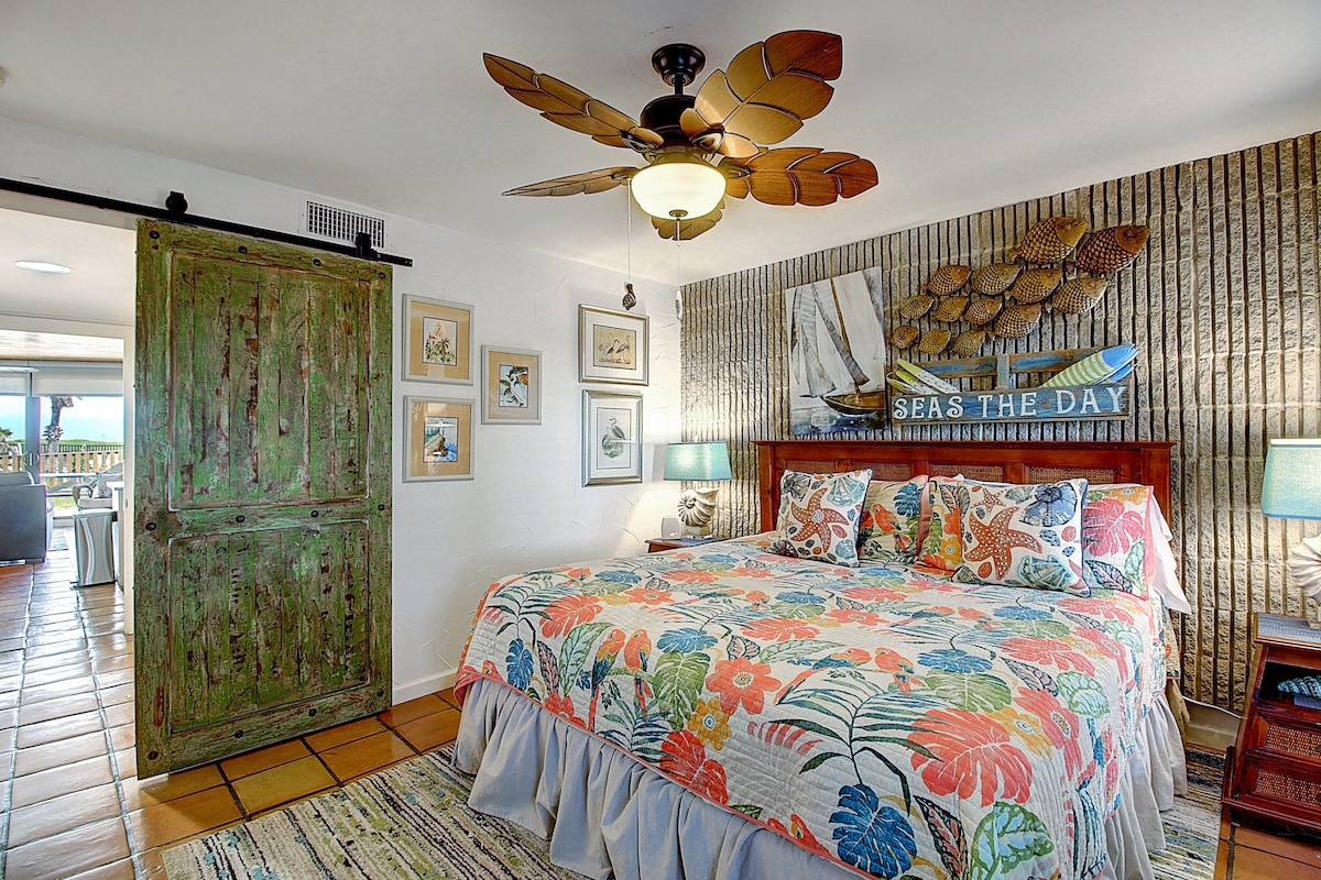 A spacious bedroom features a colorful floral quilt atop a large bed. Textured walls add character, while decorative artwork displays a nautical theme. A sliding barn door leads to another room, and two bedside lamps provide gentle illumination, creating a cozy ambiance.