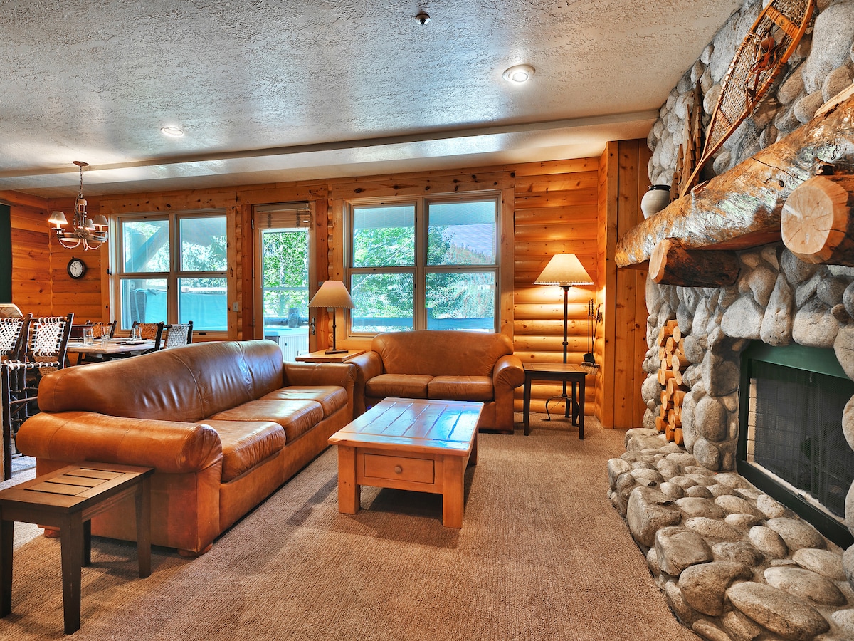 A spacious living area features a large leather sofa complemented by a smaller matching couch. A wooden coffee table sits in the center, while a stone fireplace is framed by warm wooden paneling. Natural light enters through multiple windows, enhancing the comforting ambiance.