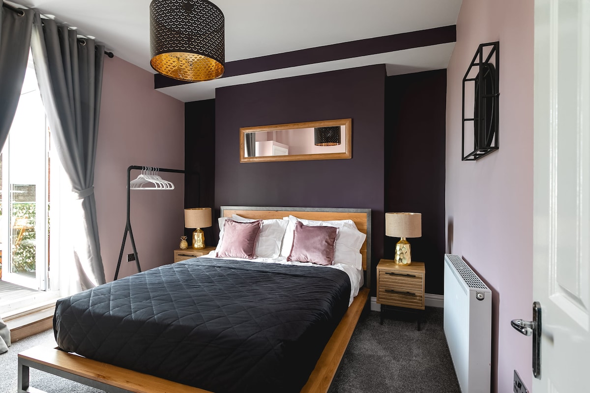 A modern bedroom showcases a spacious bed dressed in dark linens, with soft decorative pillows. Elegant bedside lamps provide a warm glow, and a large mirror reflects the room's style. Natural light enters through glass doors leading to a private balcony.