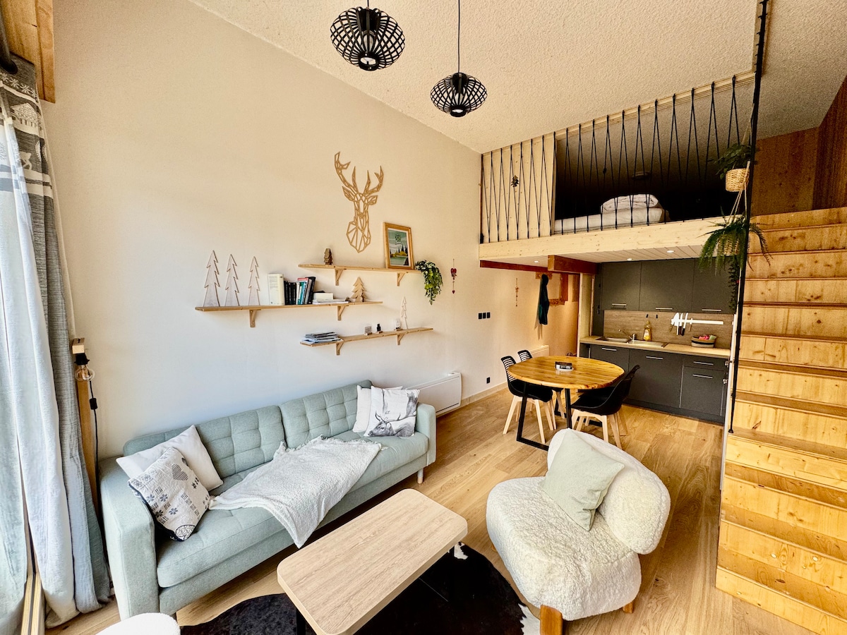 A living area features a light-colored sofa and a soft armchair, positioned near a natural wood dining table. Shelves hold decorative items, and a lofted sleeping area is visible, accessed by a wooden staircase. Large windows allow ample light to fill the space.