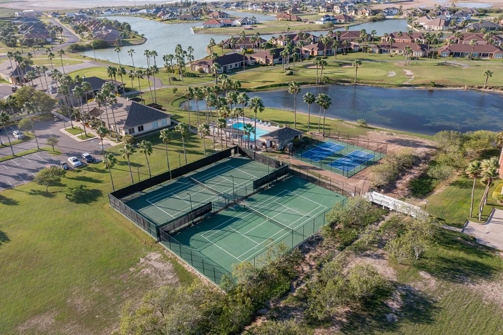 The aerial view captures a well-maintained tennis court area surrounded by palm trees and green grass. In the background, a swimming pool is visible near a clubhouse, with a serene lake and residential homes beyond, highlighting the peaceful community environment.