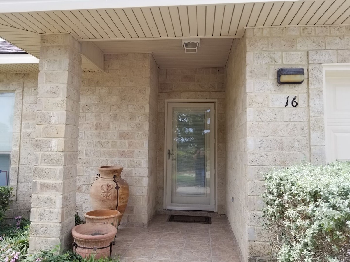 The entrance features stone exterior walls with a covered porch. A glass door is set within the stone archway, flanked by decorative terracotta pots. Green shrubs and plants surround the entrance, adding a touch of nature to the inviting facade.