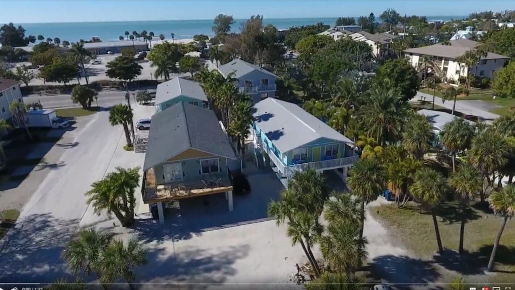 An aerial view captures the layout of Casa Coconut, showcasing two adjacent units surrounded by palm trees and greenery. The Gulf of Mexico glimmers in the background, while the nearby street provides access to local amenities and the beach.