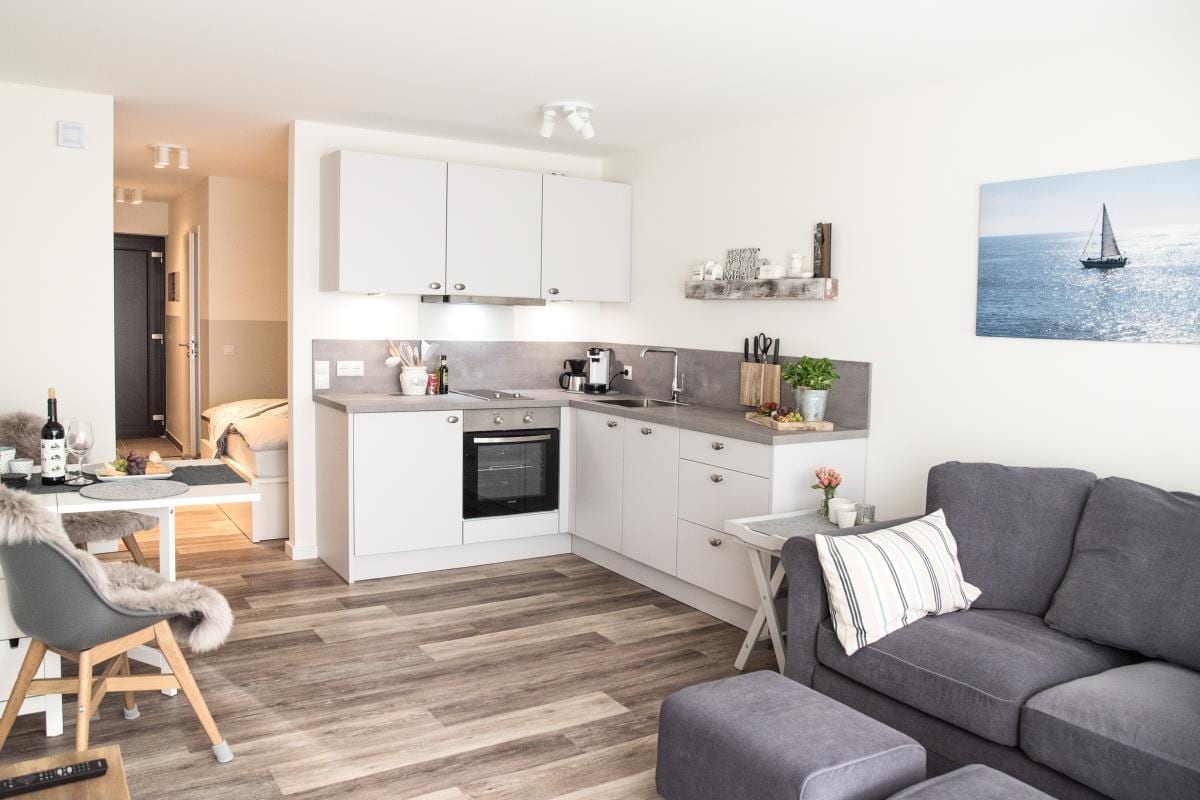 A modern kitchen area features sleek cabinetry, a built-in oven, and a countertop with small appliances. A cozy seating area is composed of a plush, gray sofa and a wooden dining table. Light wood flooring complements the neutral color palette, creating an inviting atmosphere.