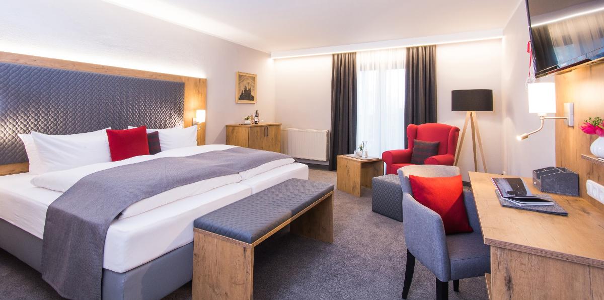 A spacious hotel room showcases a large bed with grey bedding accented by red cushions. A seating area is provided with a red armchair and a wooden desk next to a flat-screen TV. Natural light filters in through the large window, creating a bright atmosphere.