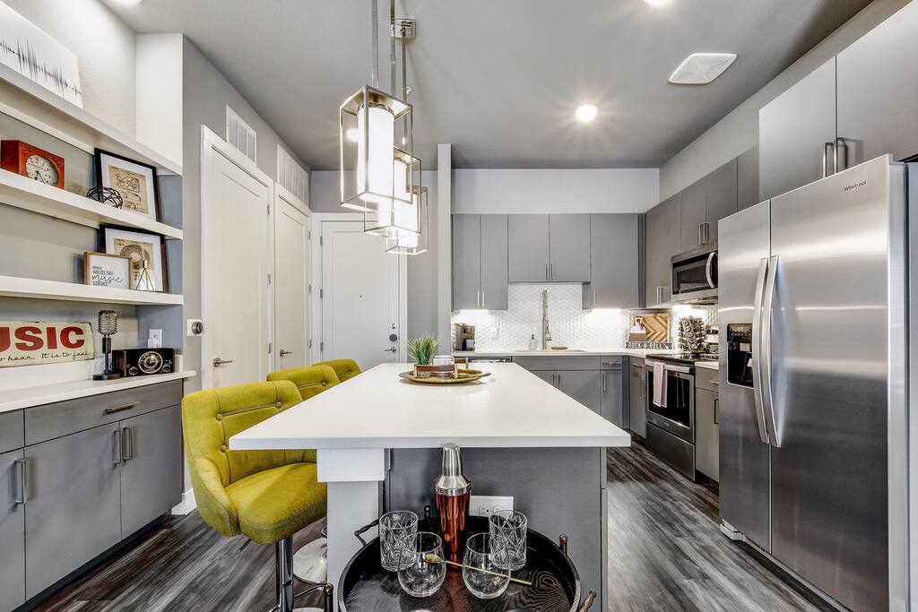 A modern kitchen showcases sleek gray cabinetry and stainless steel appliances. A central island with a white countertop provides seating with vibrant yellow bar stools. Decorative elements, including framed art and a tray of glasses, enhance the inviting atmosphere.