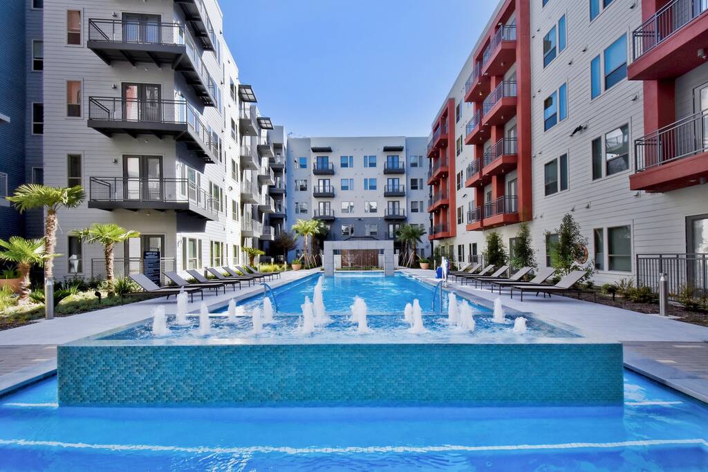 A resort-style swimming pool is centered in a landscaped courtyard, featuring a series of fountains that create a soothing water display. Sun loungers are arranged around the pool area, framed by palm trees and the modern apartment buildings that surround the space.