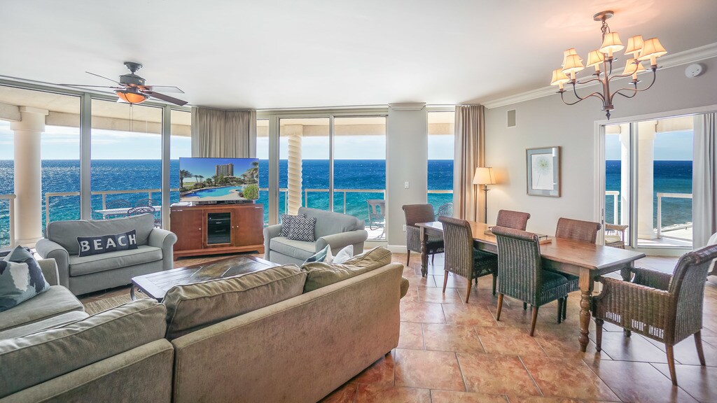 The living area provides expansive windows with stunning ocean views, adorned with comfortable seating and a flat-screen television. A dining table accommodates six, complemented by additional seating nearby. Warm lighting is complemented by ceiling fans, enhancing the relaxed coastal ambiance.