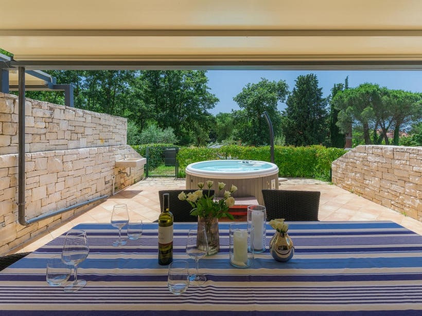A dining table is set on a covered terrace, featuring striped tablecloth and glasses. A bottle of wine and a floral arrangement enhance the setting. In the background, a jacuzzi invites relaxation, surrounded by a green garden and stone walls.