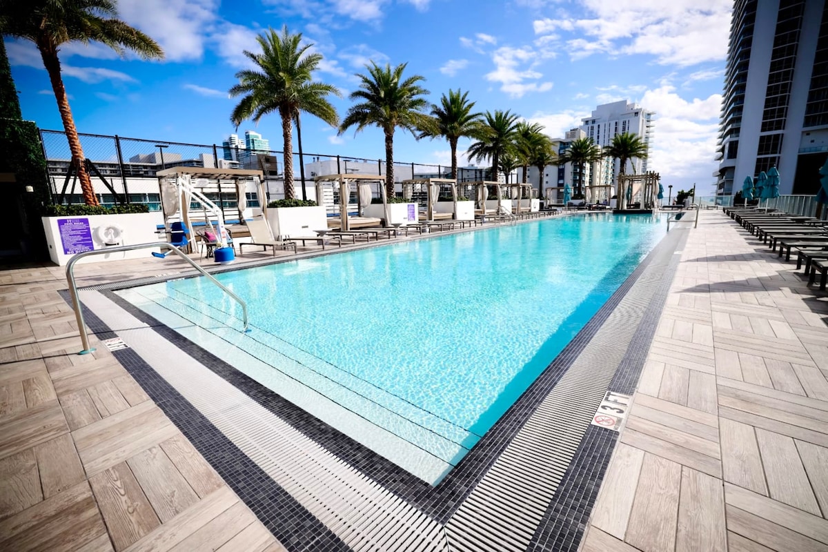 An inviting pool area is highlighted by clear blue waters and surrounded by palm trees. Sun loungers are arranged along the poolside, offering ample space for relaxation. The modern deck features a textured surface and easy access steps into the water, providing a tranquil escape.