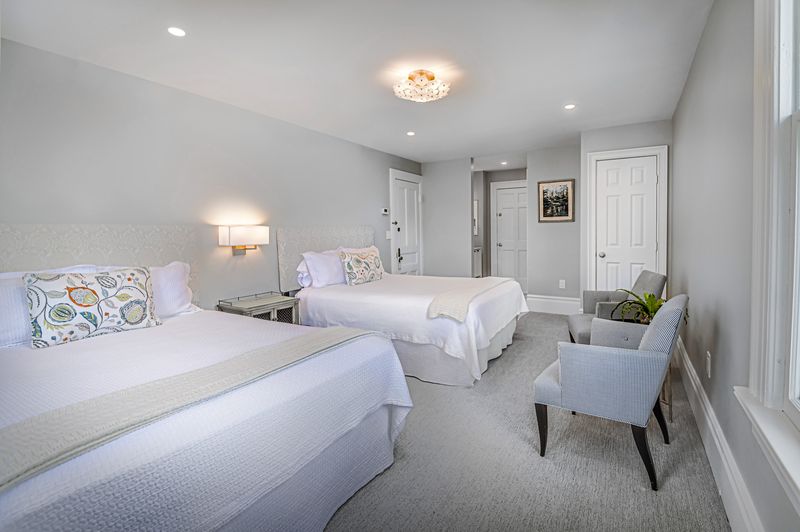 The guest room features two spacious beds with soft bedding, complemented by a modern light fixture above. A decorative chair is positioned beside a nightstand. Natural light enters through a window, illuminating the neutral-toned walls and carpet.
