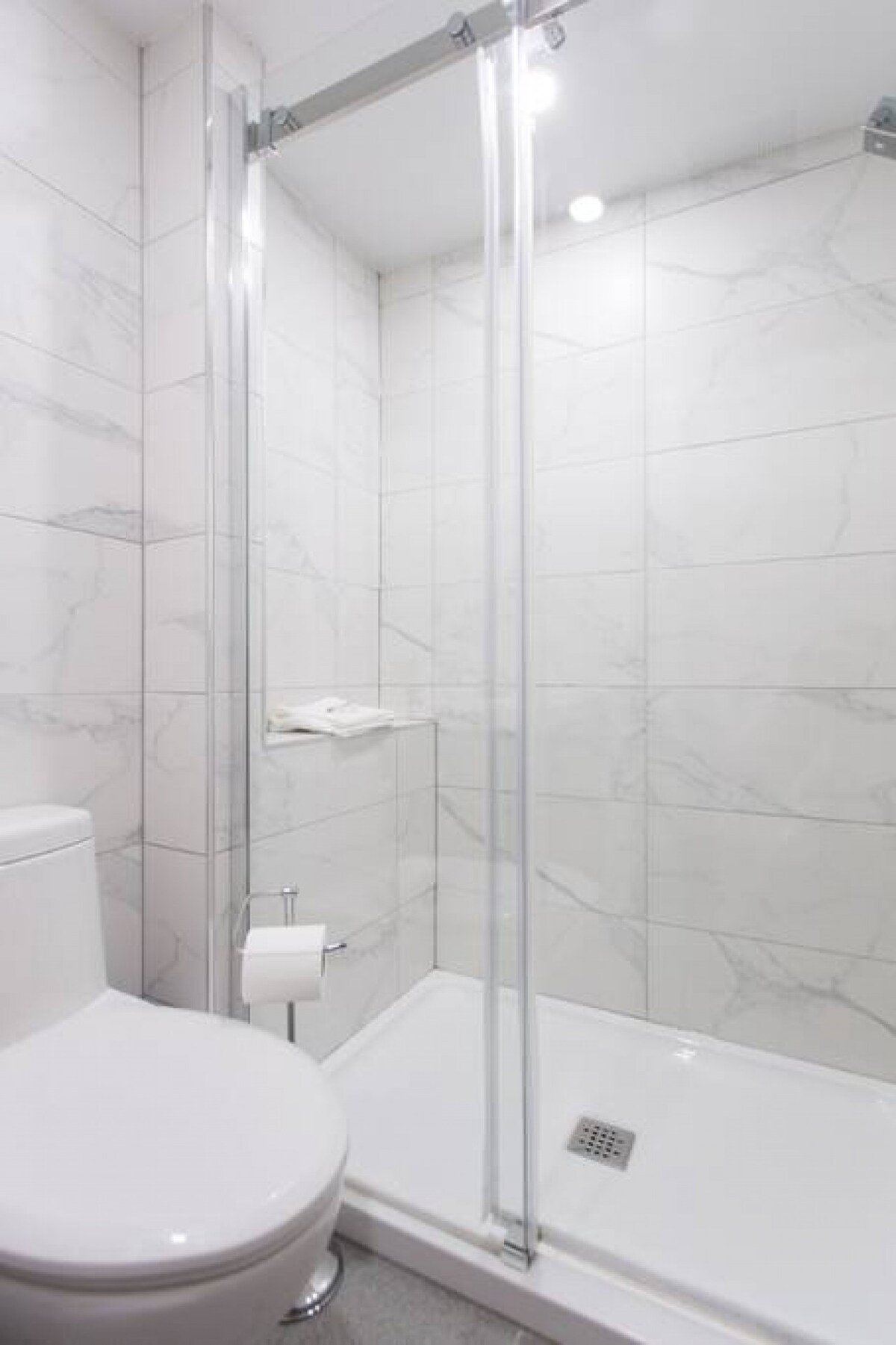 A modern bathroom features a walk-in shower enclosed by clear glass. Marble-patterned tiles cover the walls, creating a sleek aesthetic. A toilet is positioned beside the shower, with a small shelf designed for toiletries. Natural light is provided by overhead fixtures.