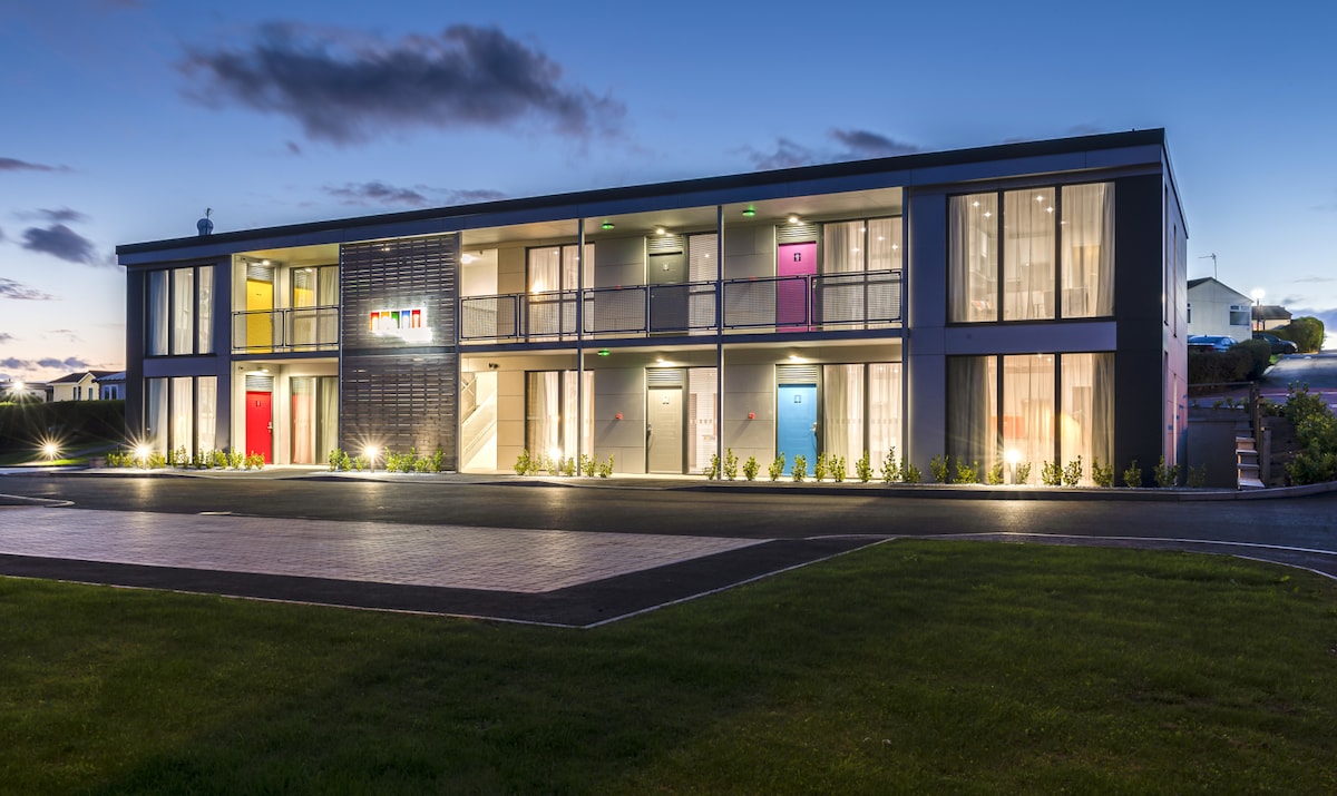 A modern two-story building is shown at dusk, featuring multiple doors in vibrant colors. Large windows enhance the exterior, allowing glimpses of the interior lighting. Well-maintained greenery lines the pathway leading to the entrance, contributing to the inviting atmosphere.