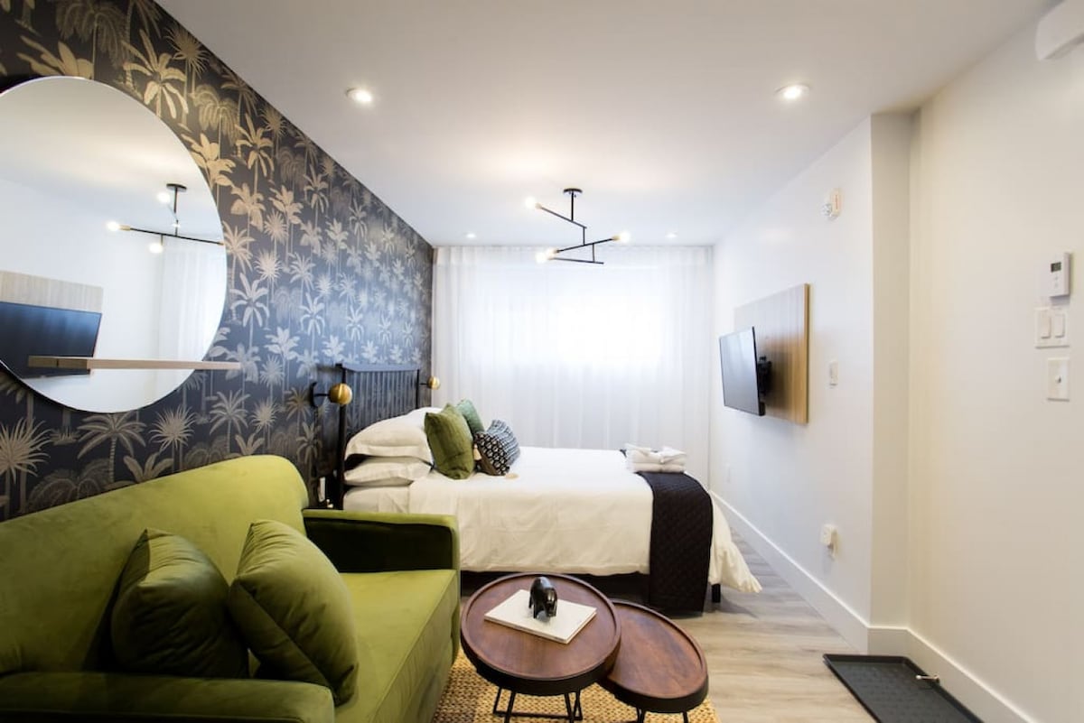 The studio features a plush green sofa opposite a spacious bed, with decorative pillows adding color. A round mirror hangs on a patterned accent wall, and natural light filters through sheer curtains. A coffee table with a small decorative item is positioned in the center.