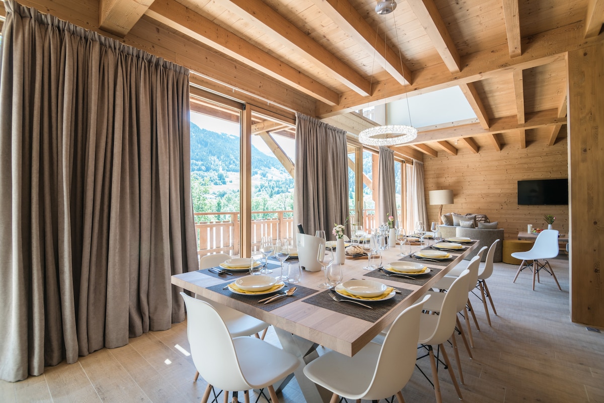A spacious dining area features a large wooden table set for ten, complemented by stylish white chairs. Natural light floods the room through multiple large windows, offering views of the surrounding mountains. Warm wooden beams and elegant drapes enhance the intimate ambiance.