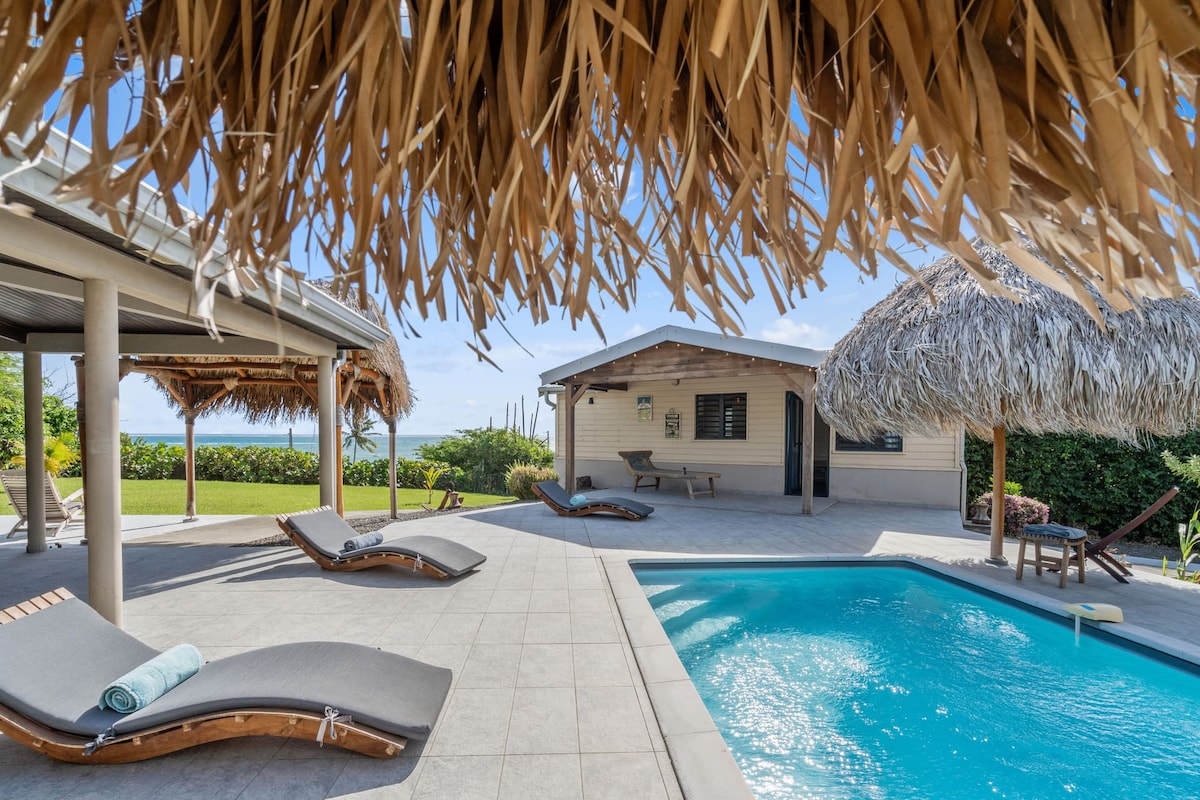 A spacious outdoor area features a refreshing pool surrounded by lounge chairs. Palm-thatched roofs provide shade, and a cozy bungalow is visible in the background. Lush greenery outlines the space, enhancing the inviting atmosphere for relaxation and leisure.