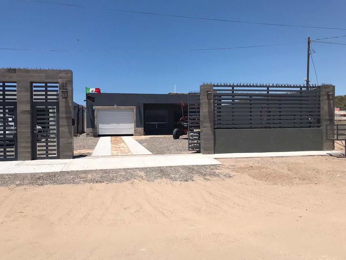 A gated entrance leads to a modern home, featuring a gray building with sleek lines and large garage doors. The pathway, lined with gravel, extends toward the structure, while a flag is visible on the property, enhancing its street appeal.